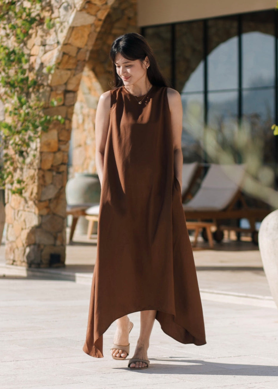 Woman wearing a brown dress standing outdoors near a stone wall and chairs.