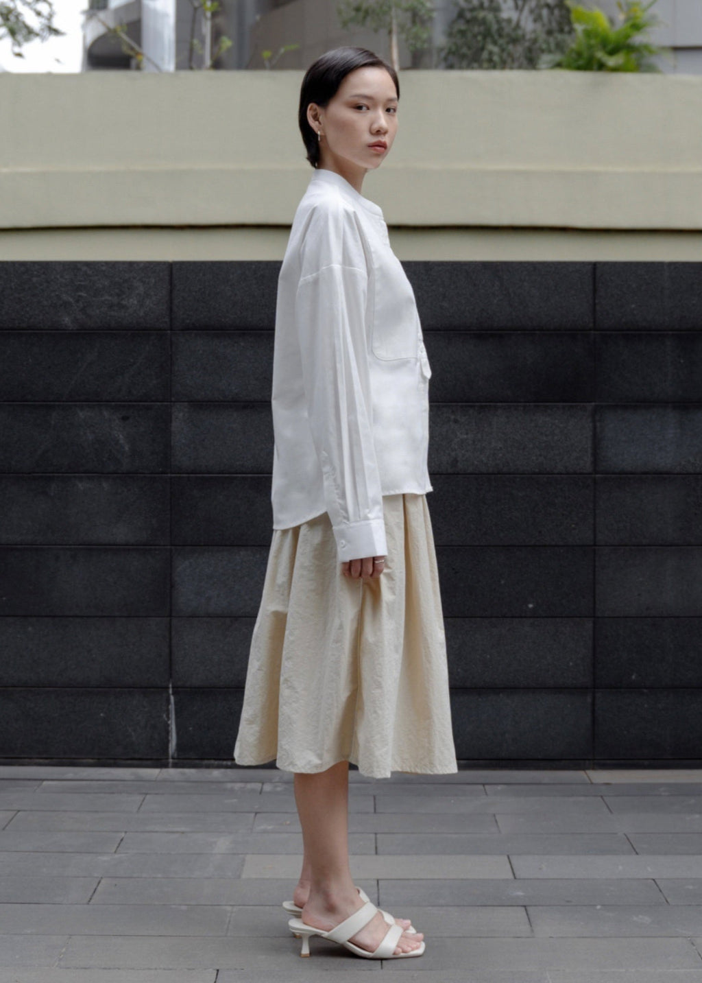 Woman wearing a white blouse and beige skirt standing against a black brick wall.