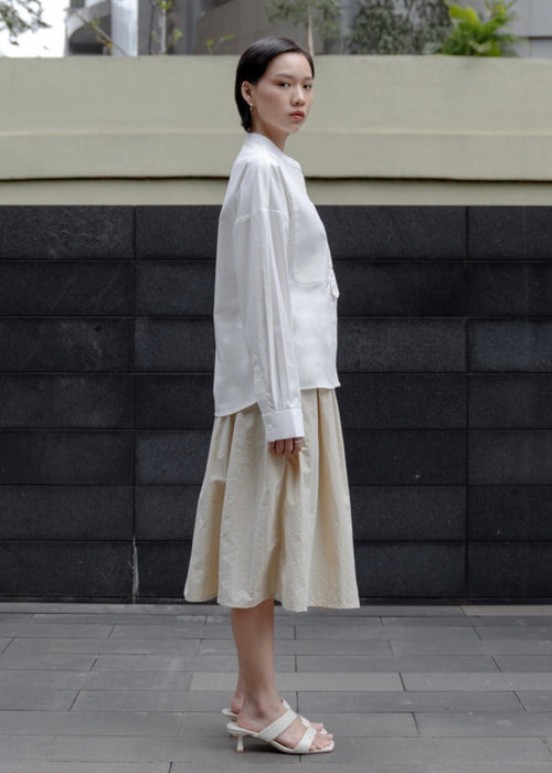 Woman wearing a white blouse and beige skirt standing against a black brick wall.