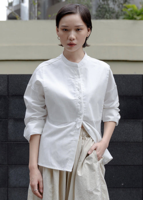 Woman wearing a white blouse and beige pants standing against a black and gray wall.