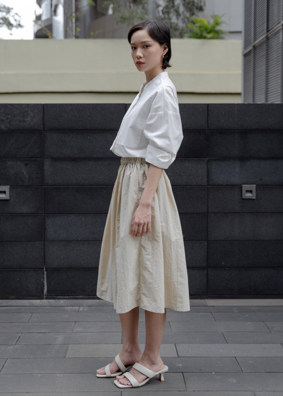 Woman in a white blouse and beige skirt standing against a black wall.