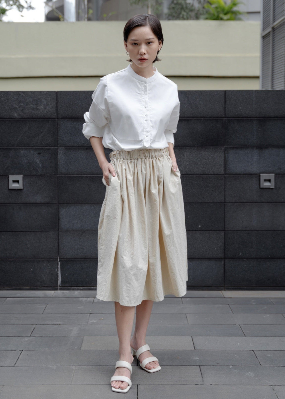 Woman wearing a white blouse and beige skirt standing against a black and gray wall.