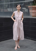 Woman in a light pink dress standing in an outdoor setting with a reflective surface and plants.