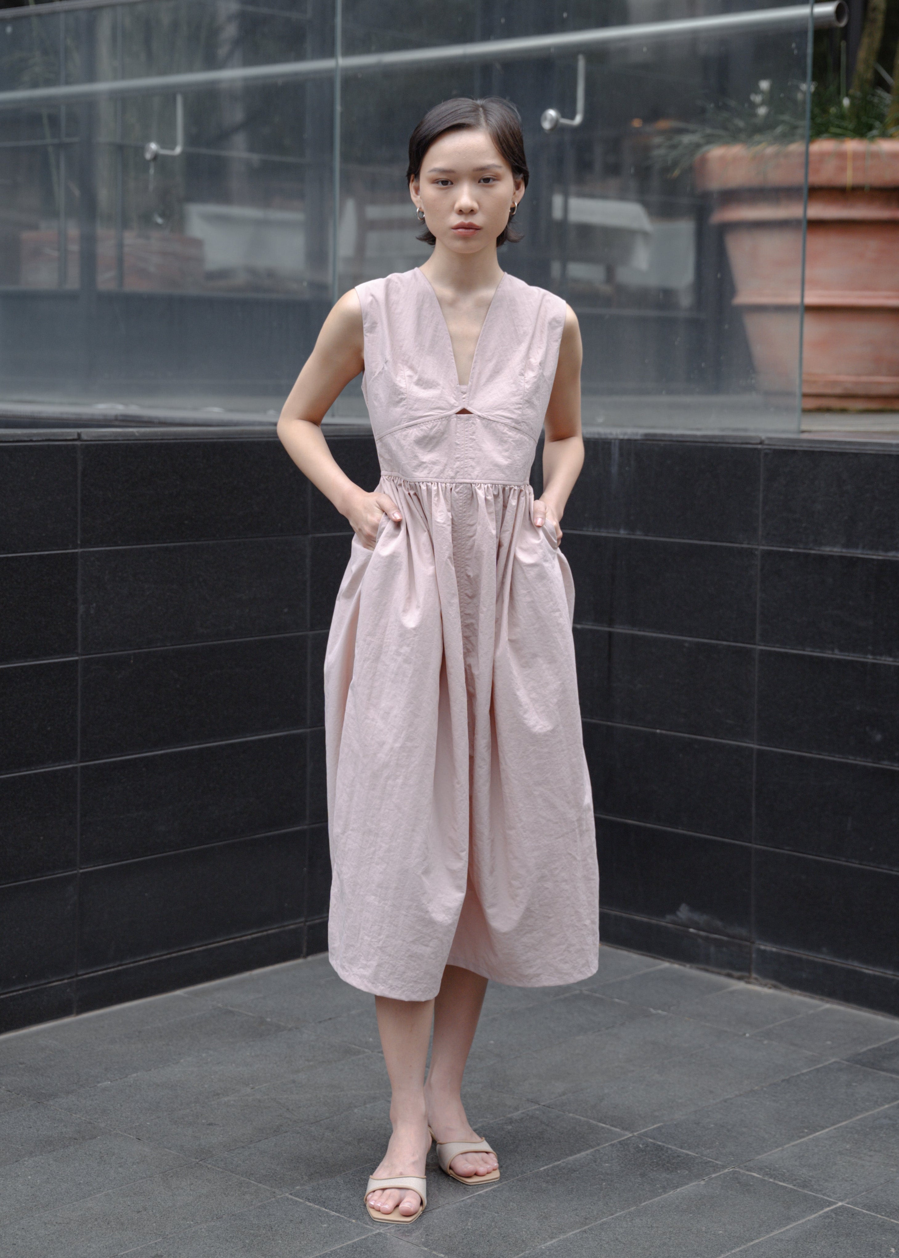Woman in a light pink dress standing in an outdoor setting with a reflective surface and plants.