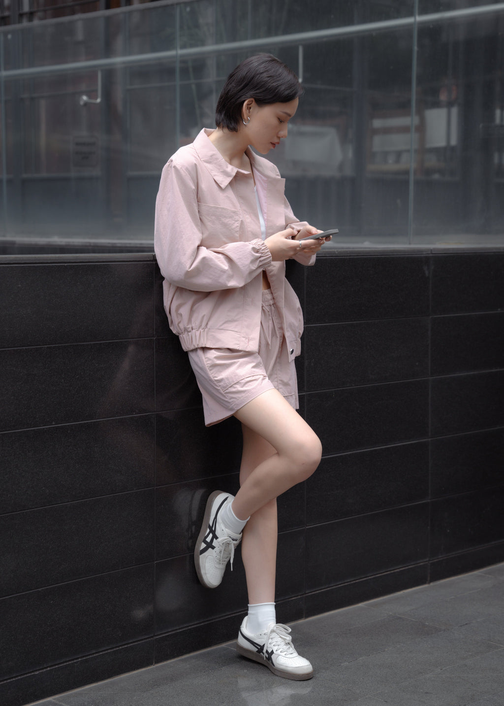 Woman in a light pink outfit sitting against a black wall, looking at her phone.