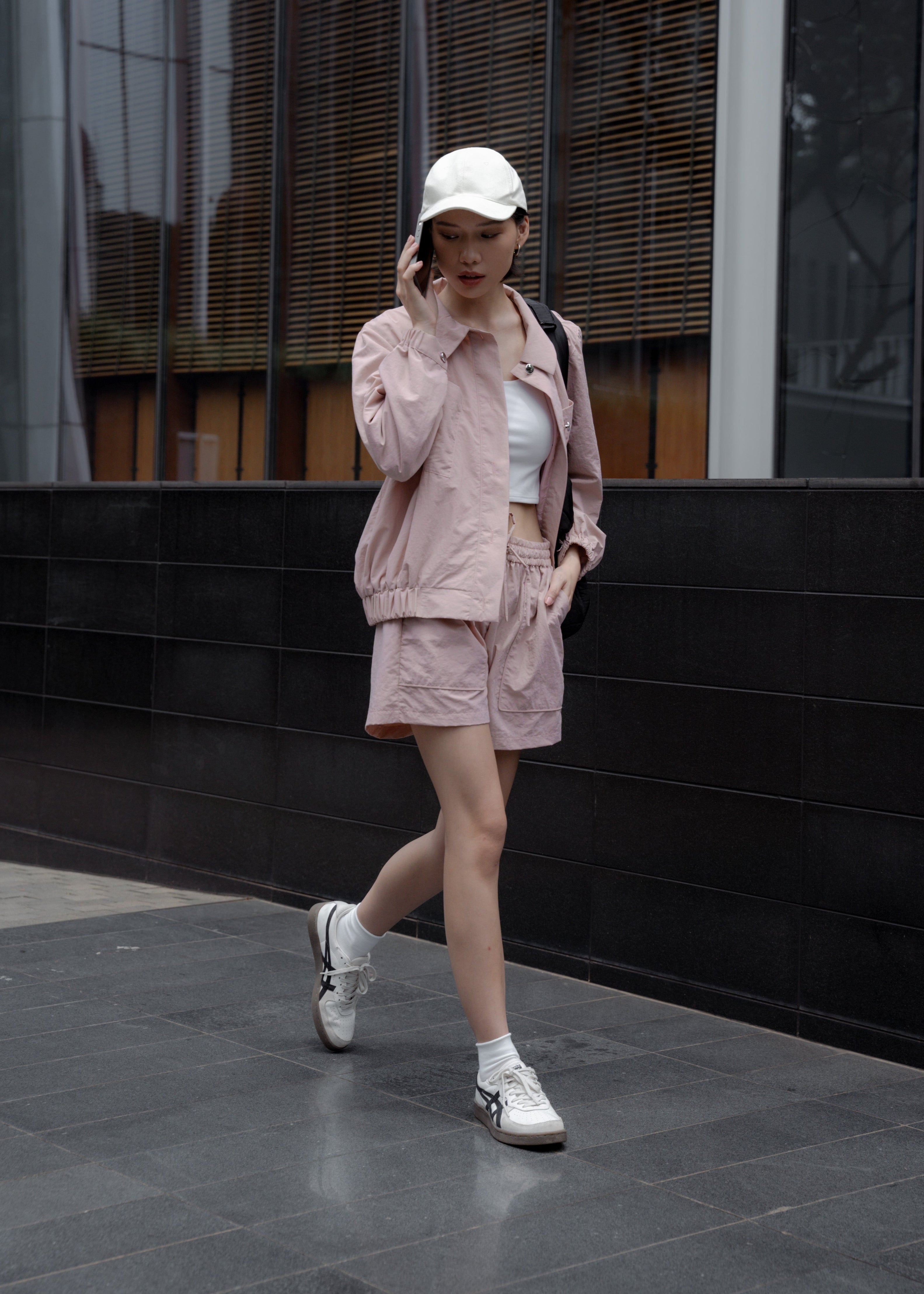 Woman in a pink jacket and white sneakers walking in front of a modern building.