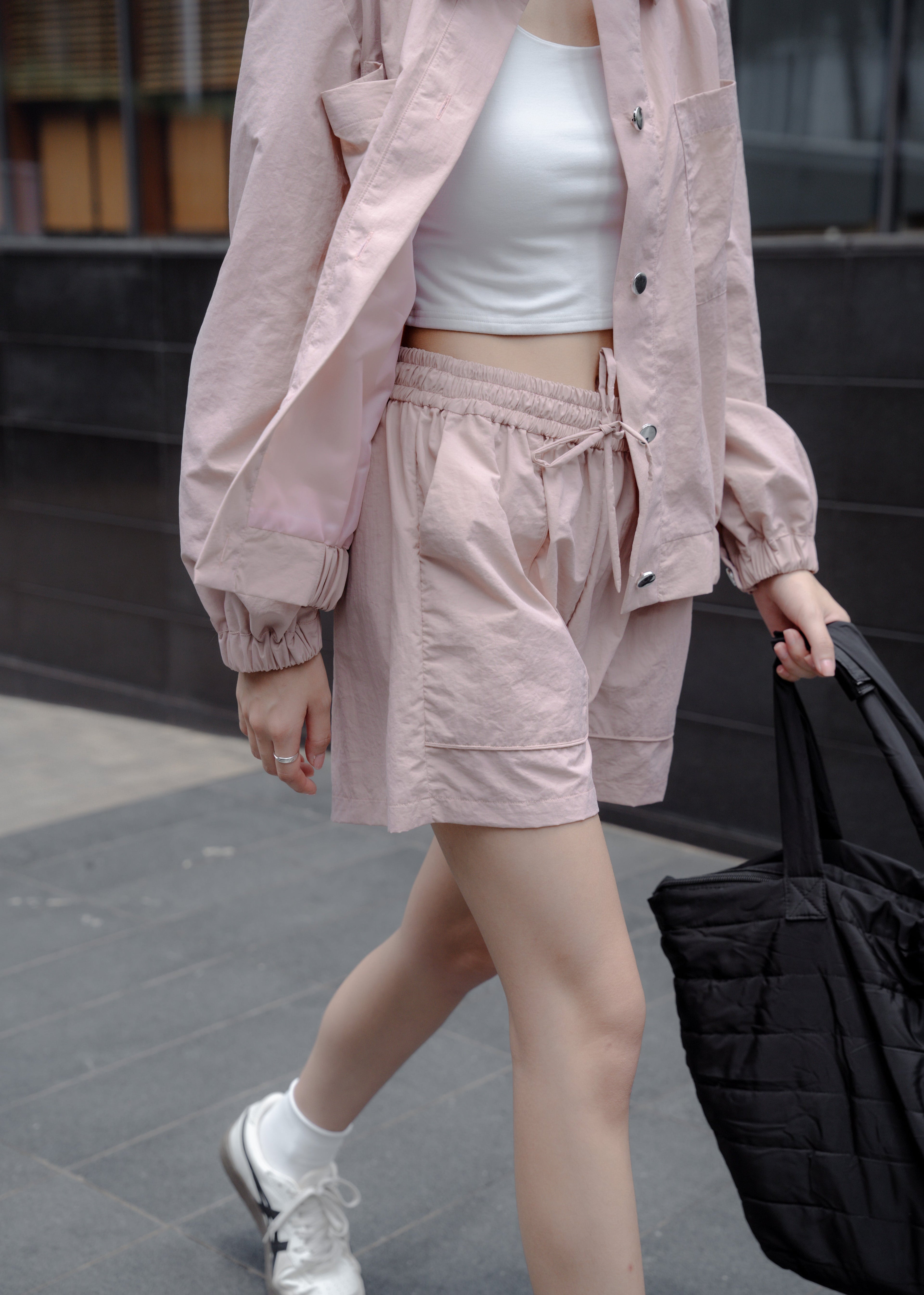 Person wearing a pink outfit with shorts and jacket, holding a black bag.