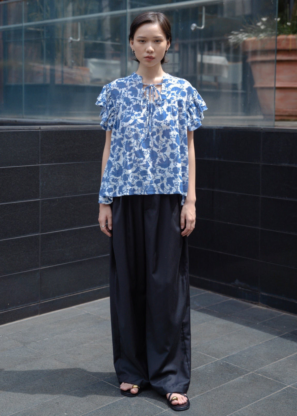 Woman wearing a blue floral blouse and black pants standing against a black tiled wall.