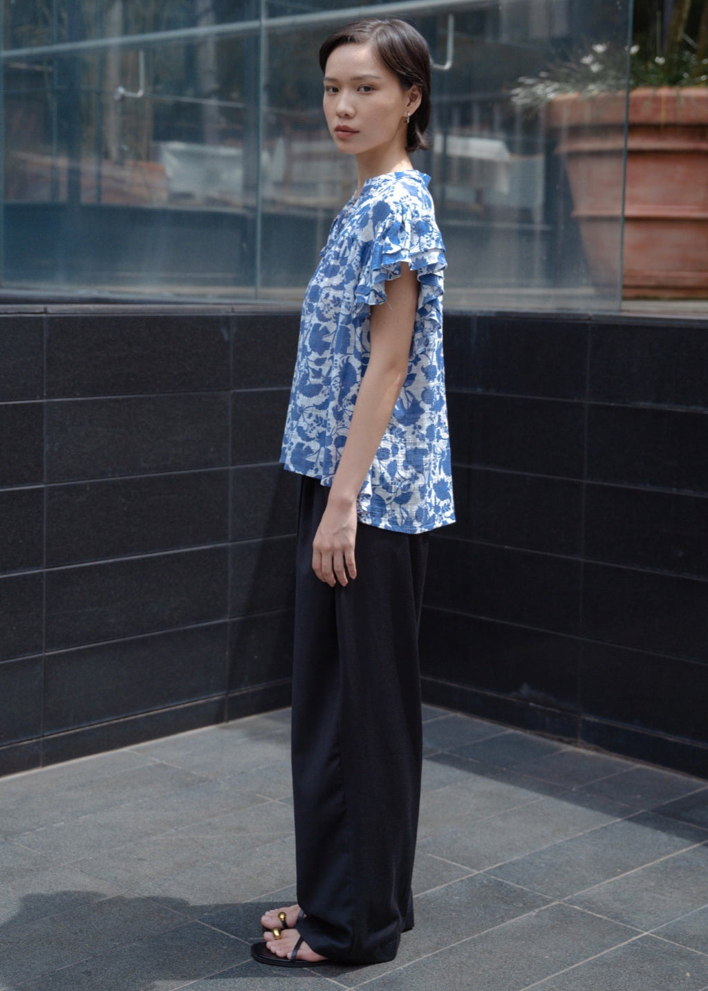 Person wearing a blue patterned shirt and black pants standing against a tiled wall.