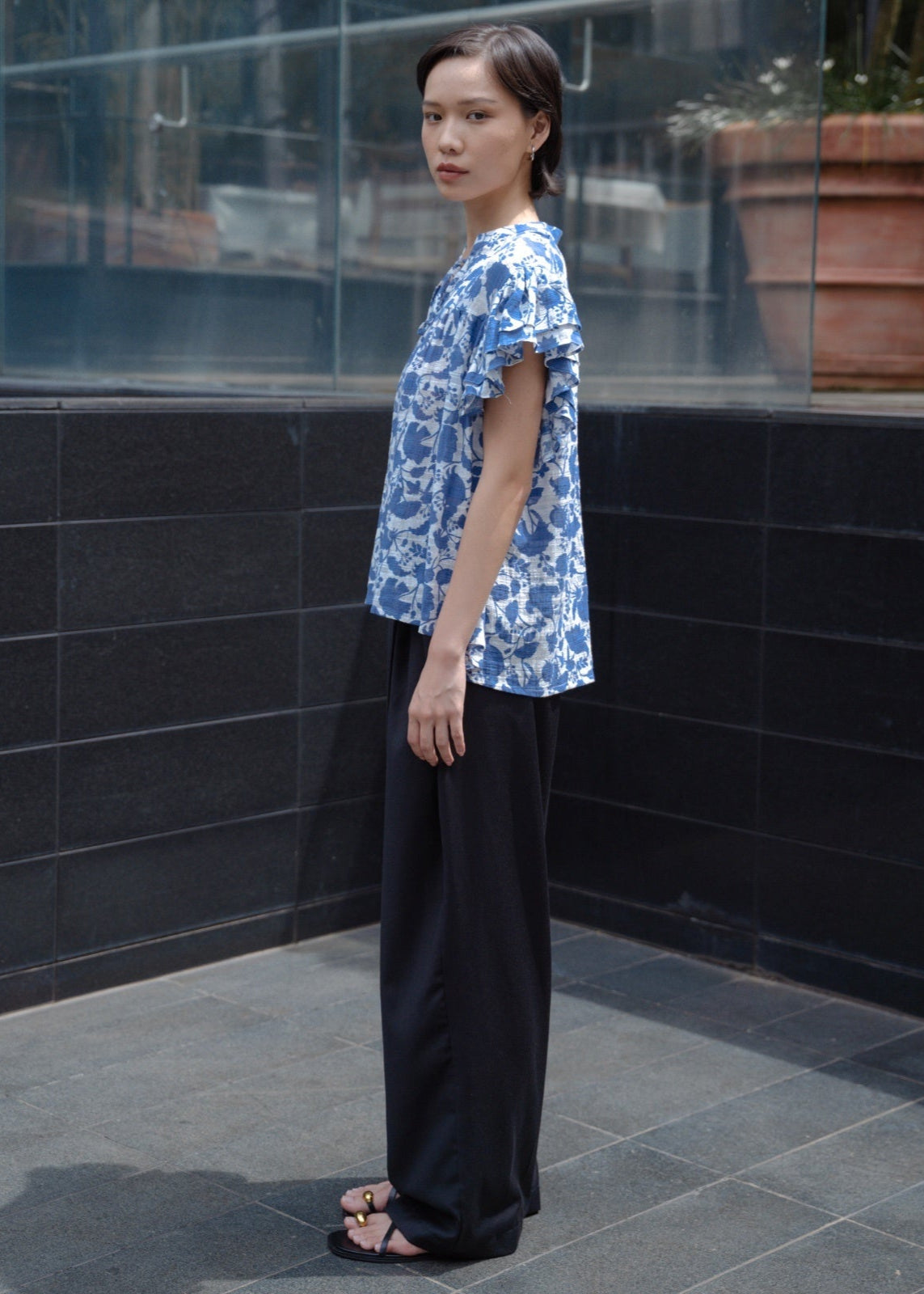 Person wearing a blue patterned shirt and black pants standing against a tiled wall.