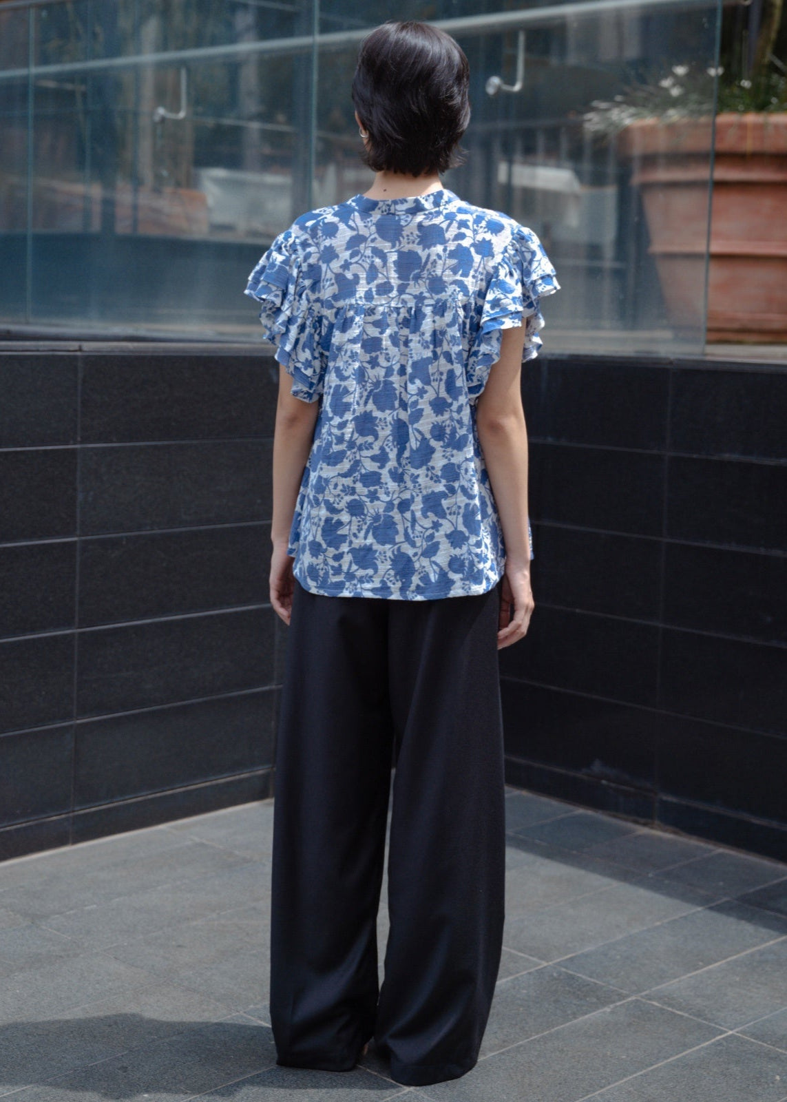 Person wearing a blue floral top and black pants standing against a reflective glass wall.