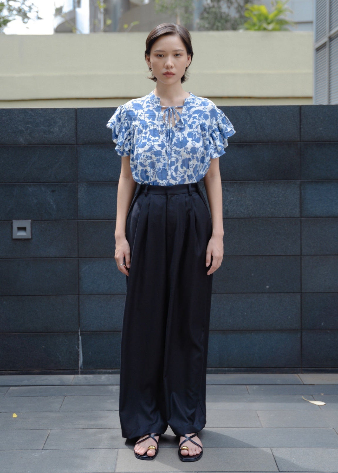 Woman wearing a blue floral top and black pants standing against a dark tiled wall.