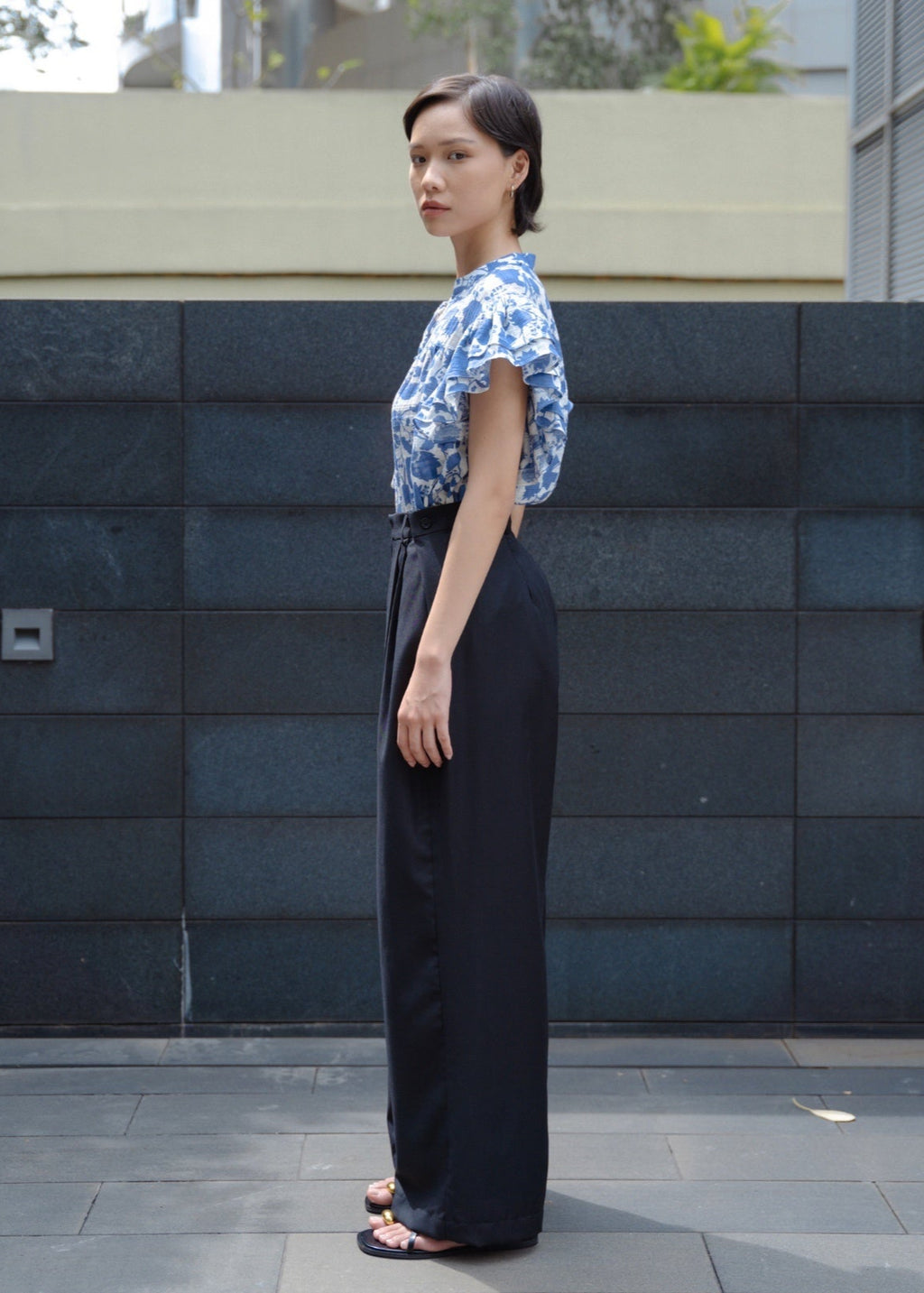 Woman wearing a blue floral top and black pants standing against a dark tiled wall.