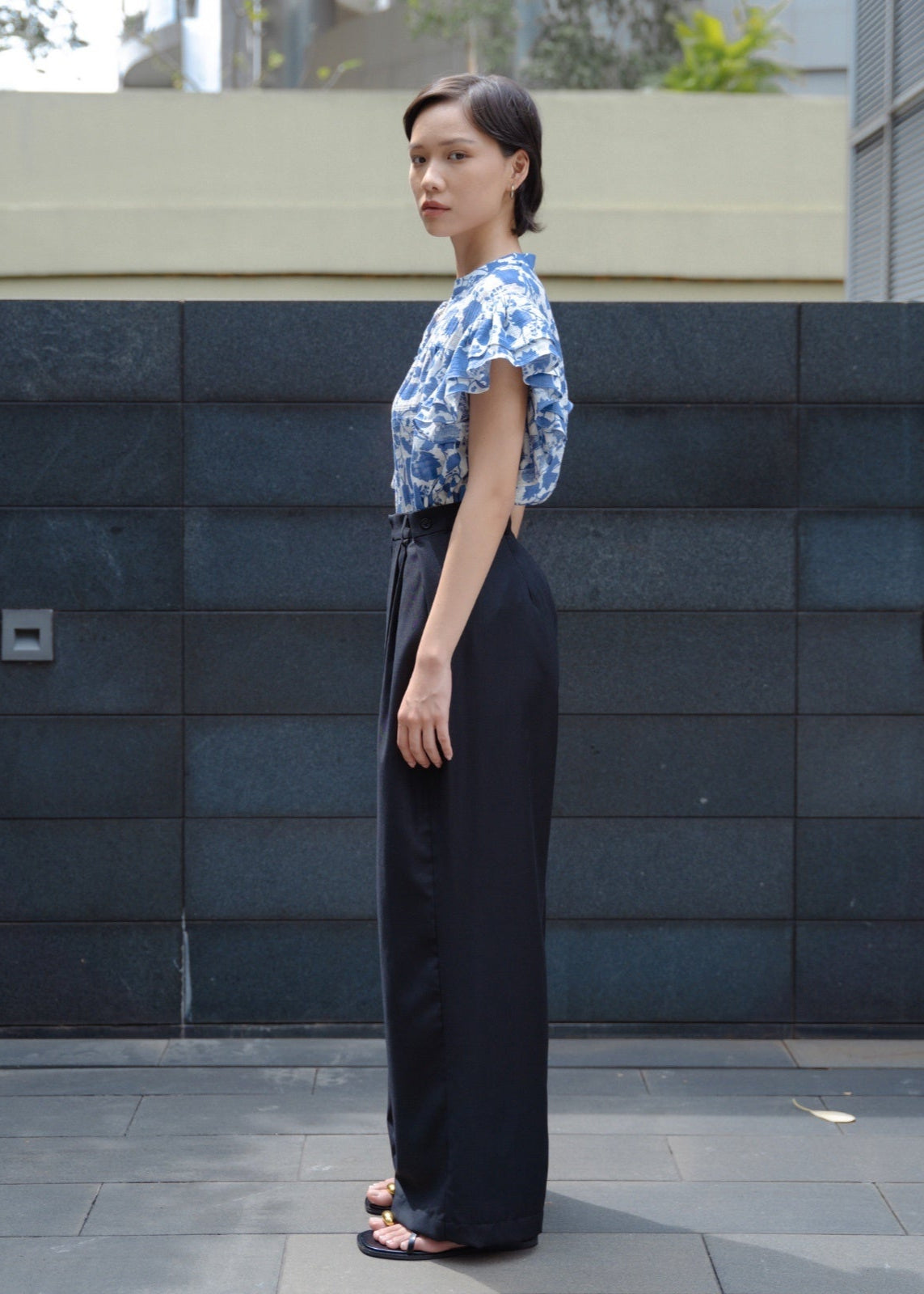 Woman wearing a blue floral top and black pants standing against a dark tiled wall.