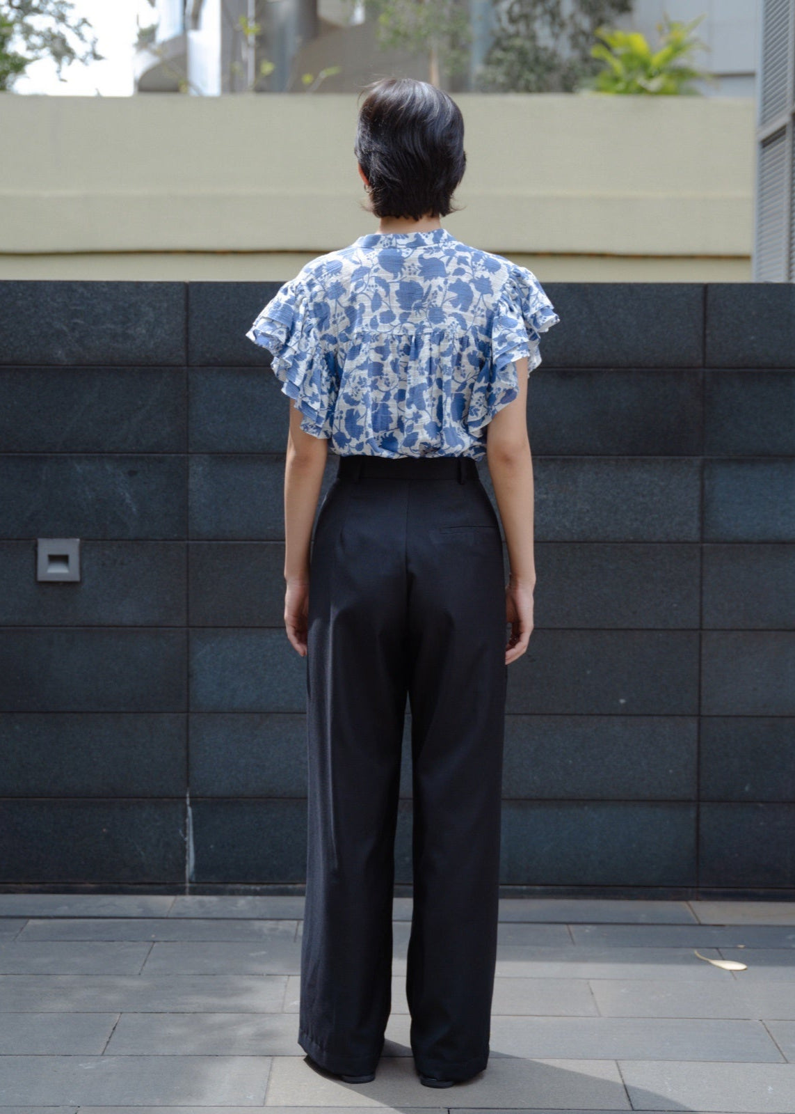 Person wearing a blue floral top and black pants standing against a dark stone wall.