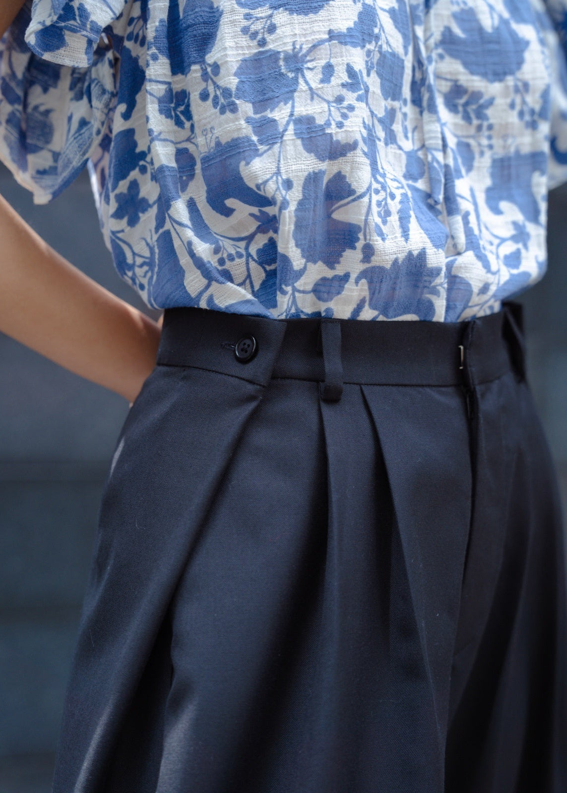 Person wearing a blue and white floral shirt with pleated navy pants against a gray background