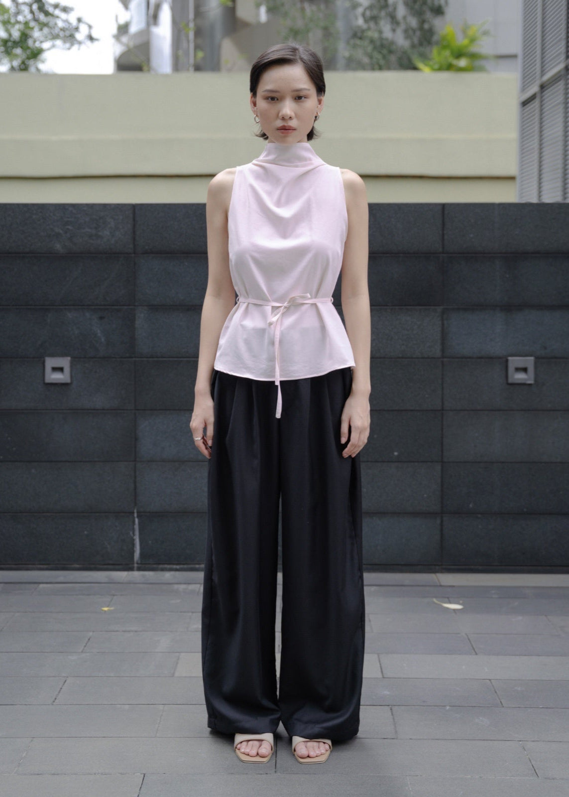 Woman wearing a light pink sleeveless top and black pants standing in front of a gray wall.