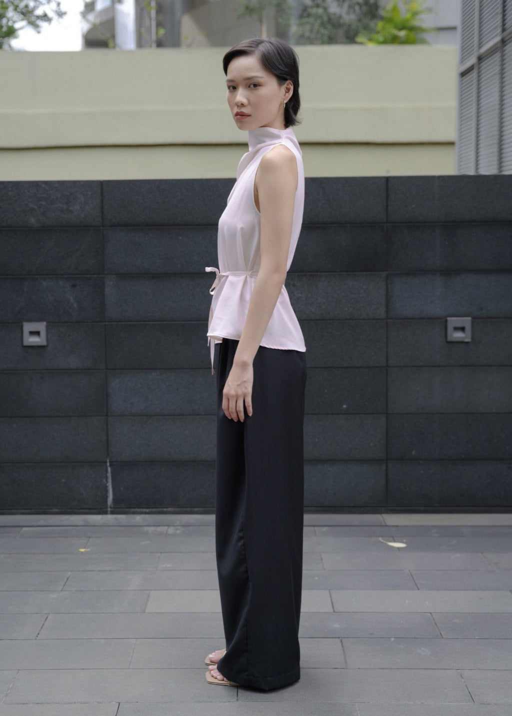 Woman wearing a white sleeveless top and black pants standing in front of a gray wall.
