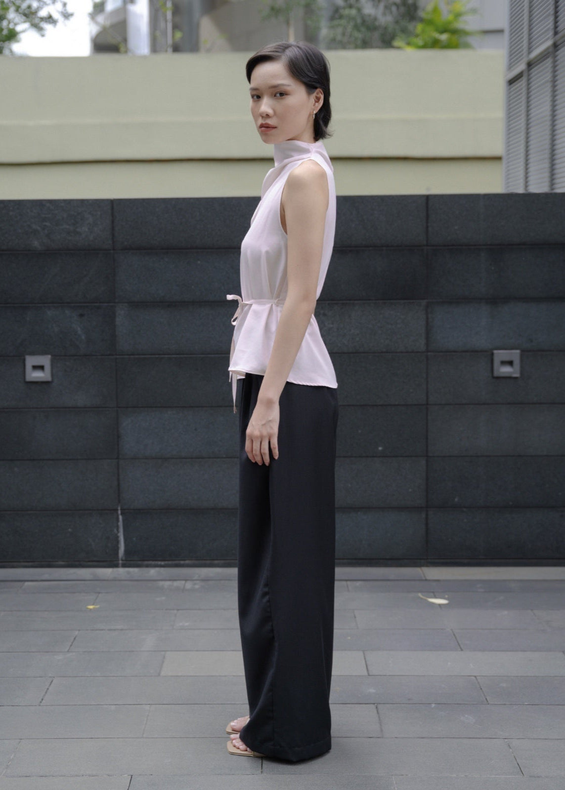 Woman wearing a white sleeveless top and black pants standing in front of a gray wall.