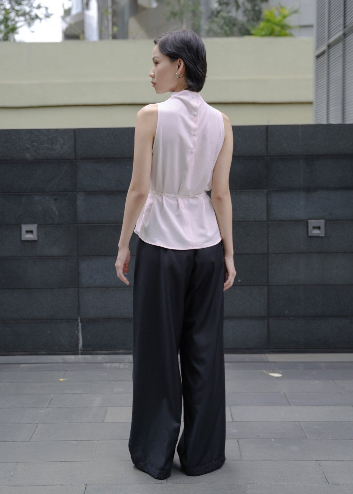 Woman wearing a sleeveless white blouse and black pants standing in front of a gray wall.