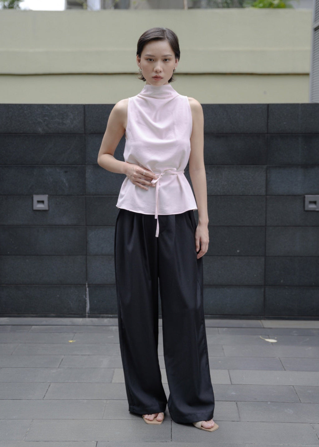 Woman wearing a light pink sleeveless top and black pants standing against a dark stone wall.