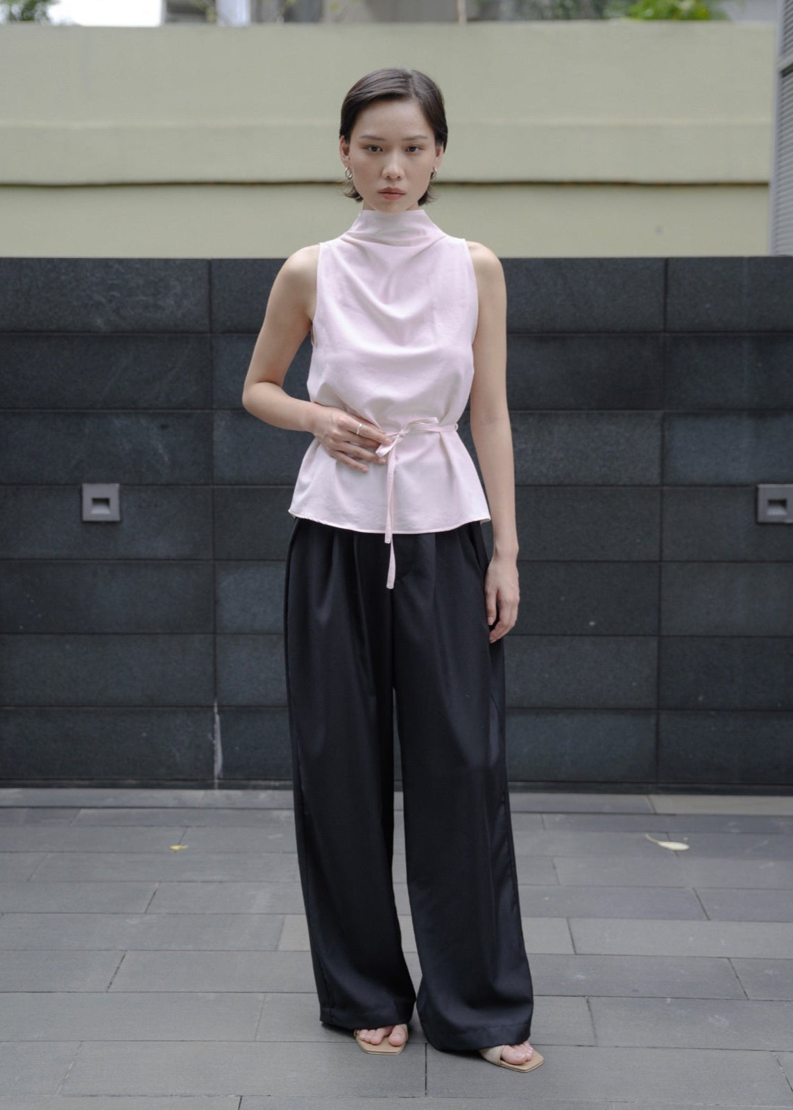 Woman wearing a light pink sleeveless top and black pants standing against a dark stone wall.
