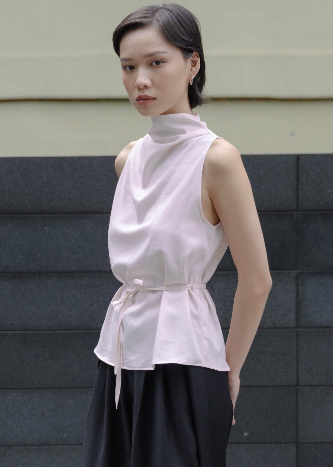 Woman wearing a light pink sleeveless top with a high collar and black pants against a gray wall.
