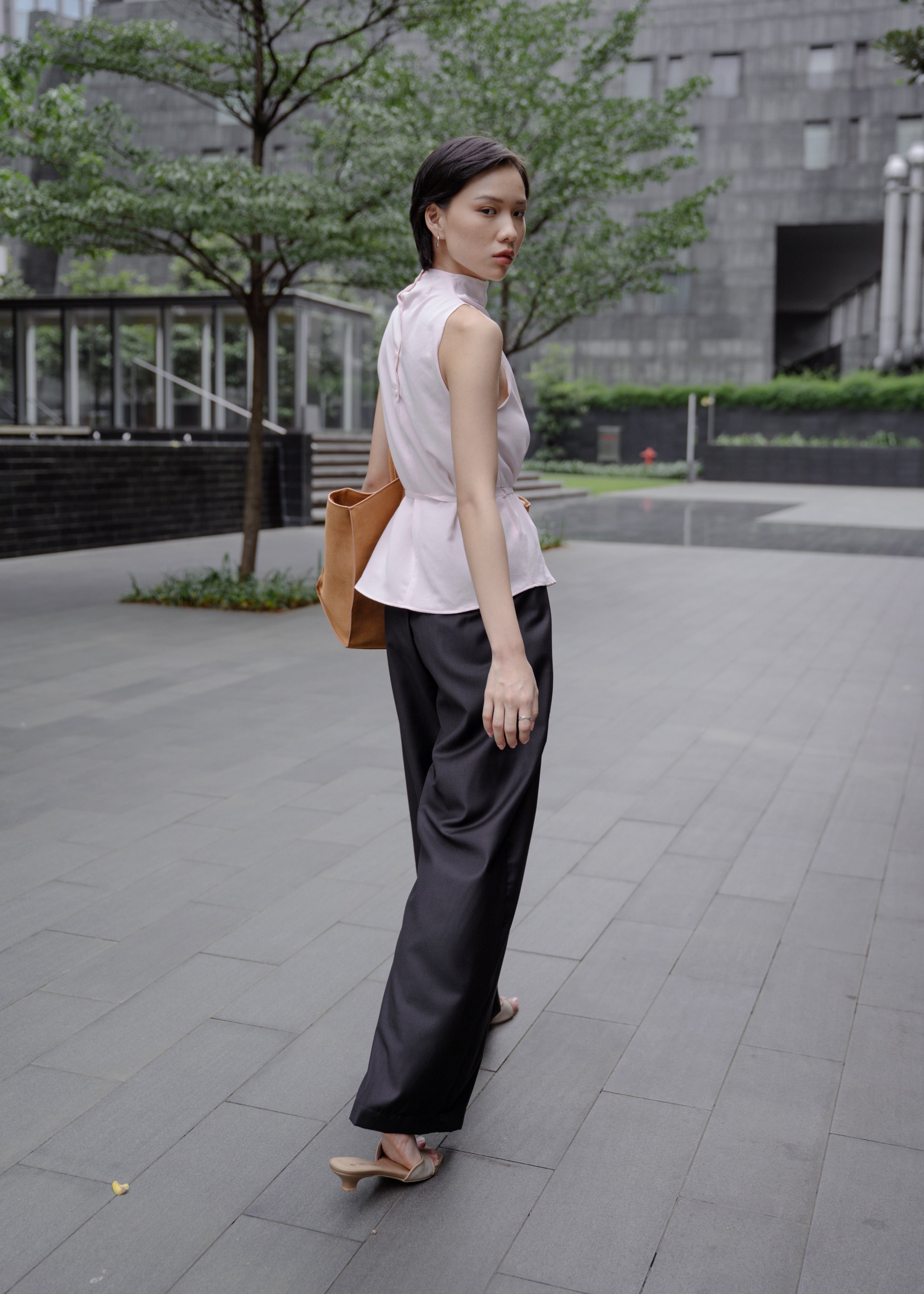 Woman walking outdoors in an urban setting with a building and trees in the background.