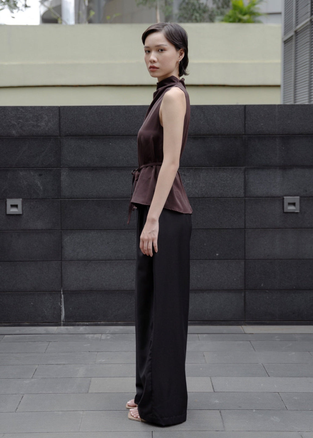 Woman in a brown sleeveless top and black pants standing against a gray wall.