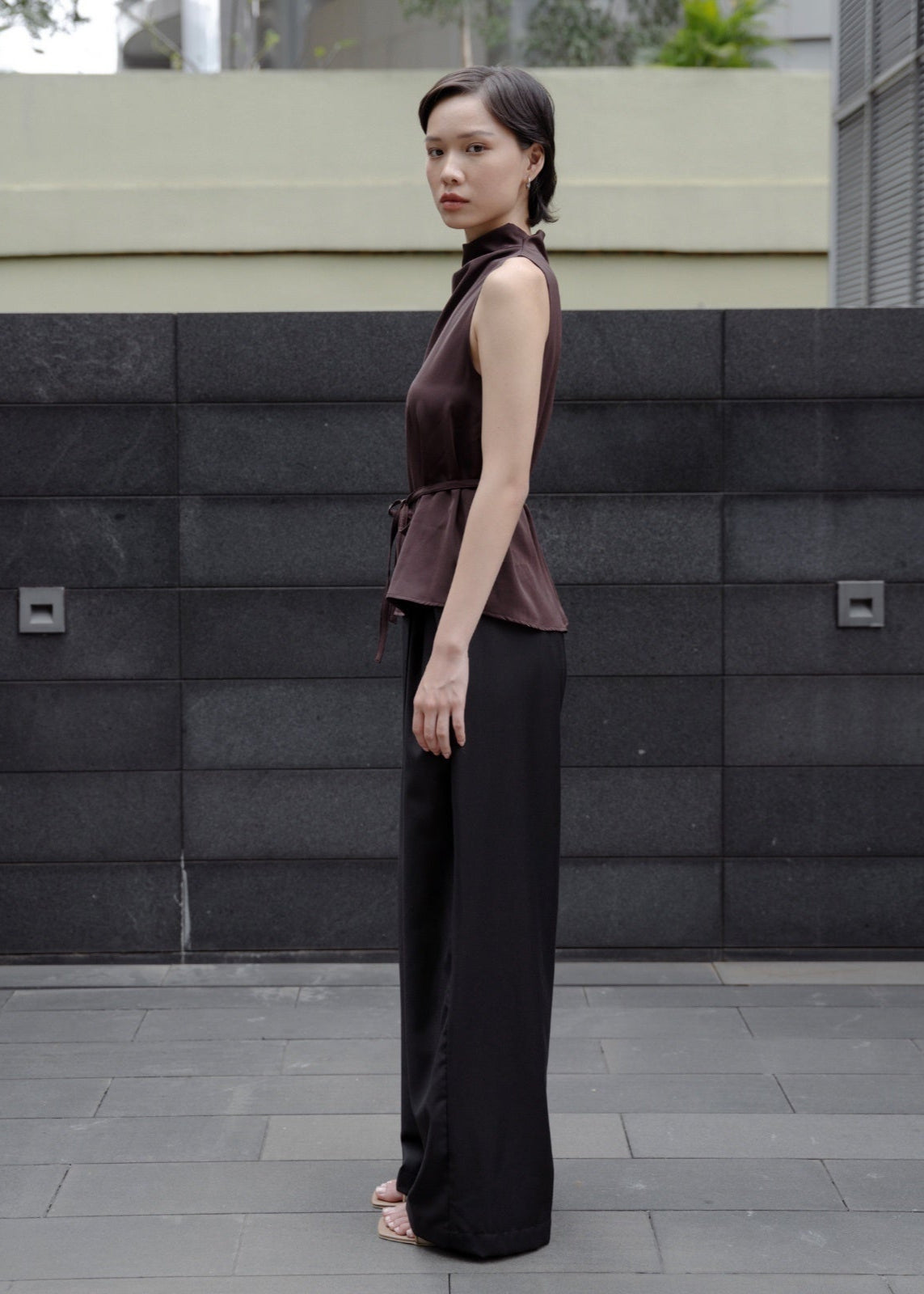 Woman in a brown sleeveless top and black pants standing against a gray wall.