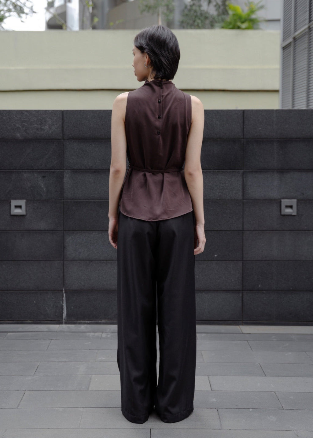 Person wearing a sleeveless brown top and black pants standing in front of a textured wall.