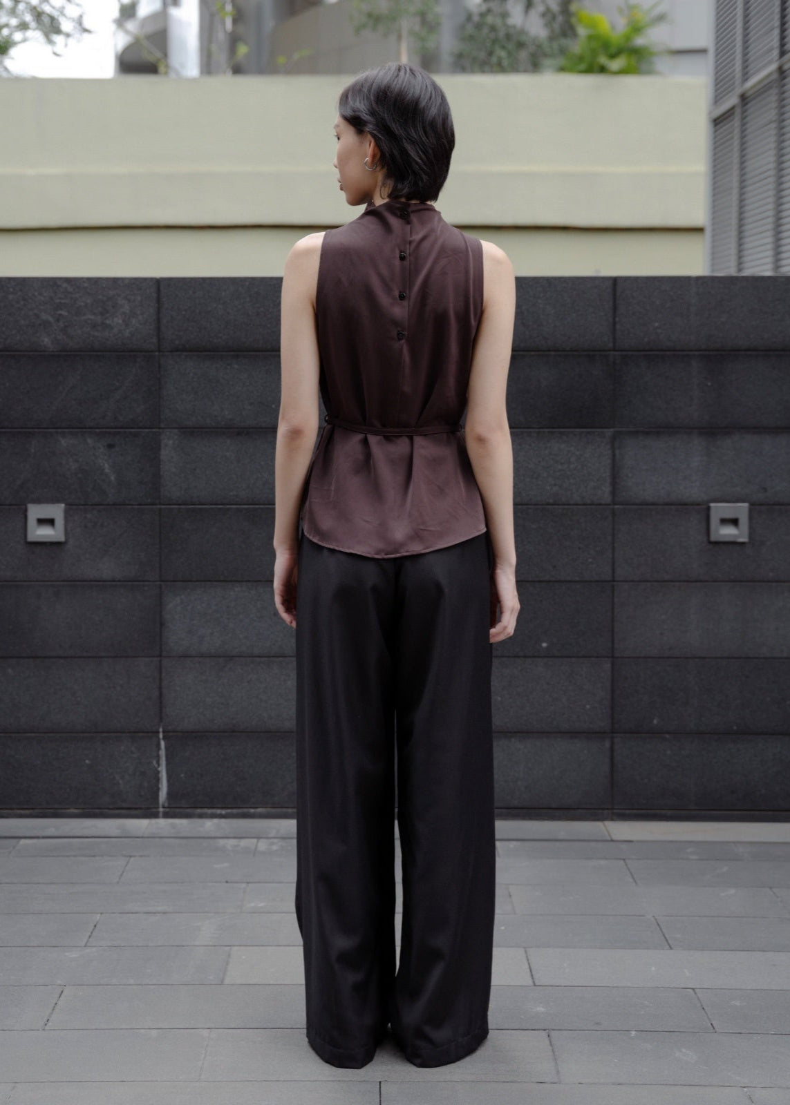 Person wearing a sleeveless brown top and black pants standing in front of a textured wall.
