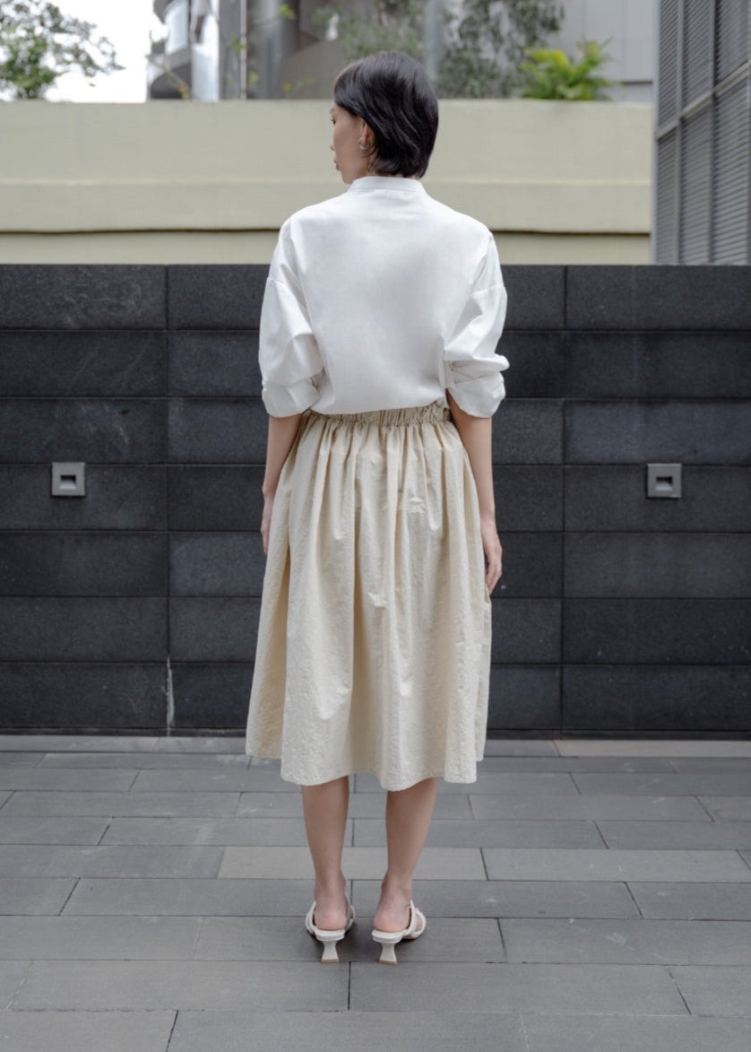 Person wearing a white shirt and beige skirt standing in front of a modern building.
