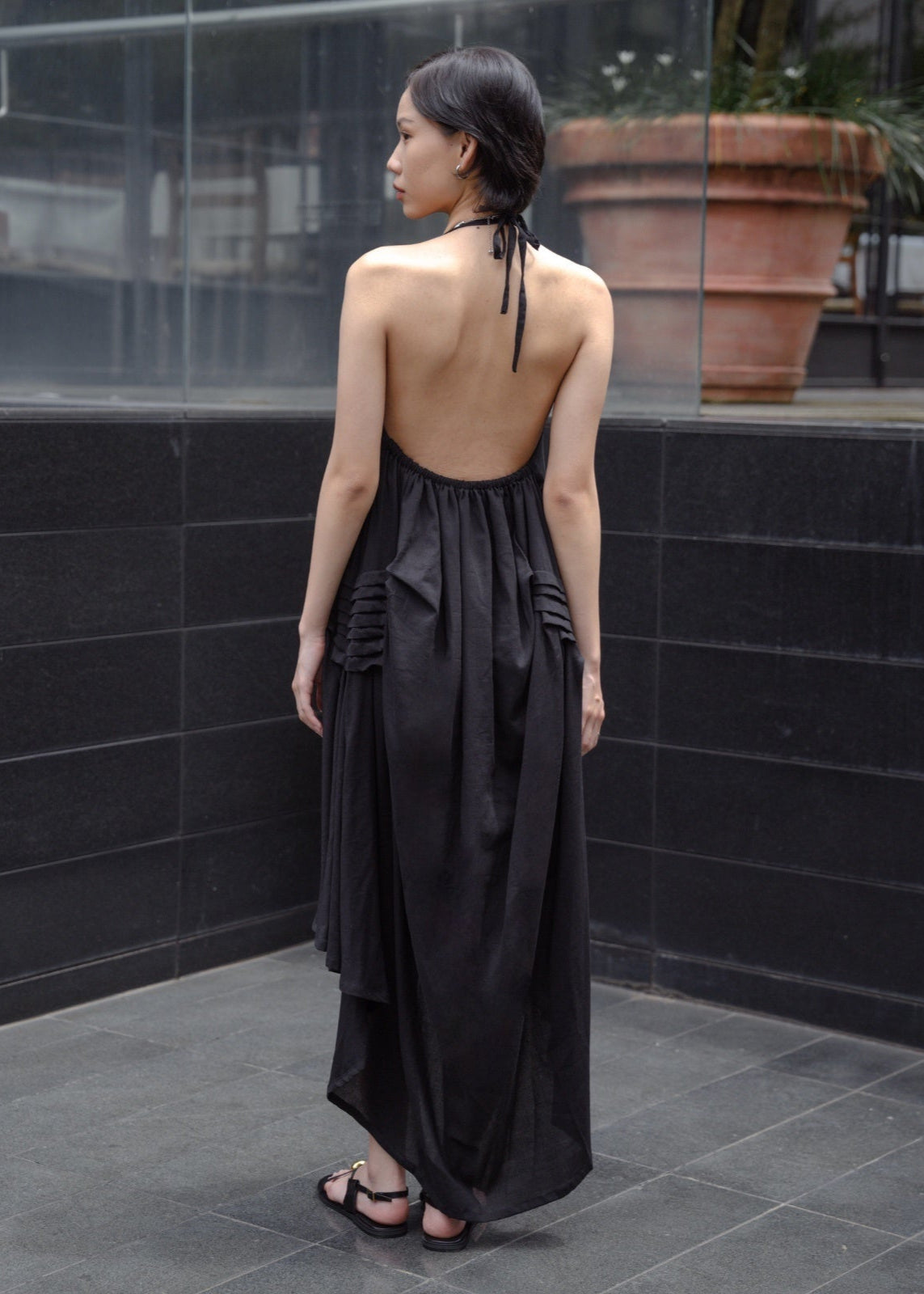 Woman wearing a black dress with an open back standing against a black tiled wall.