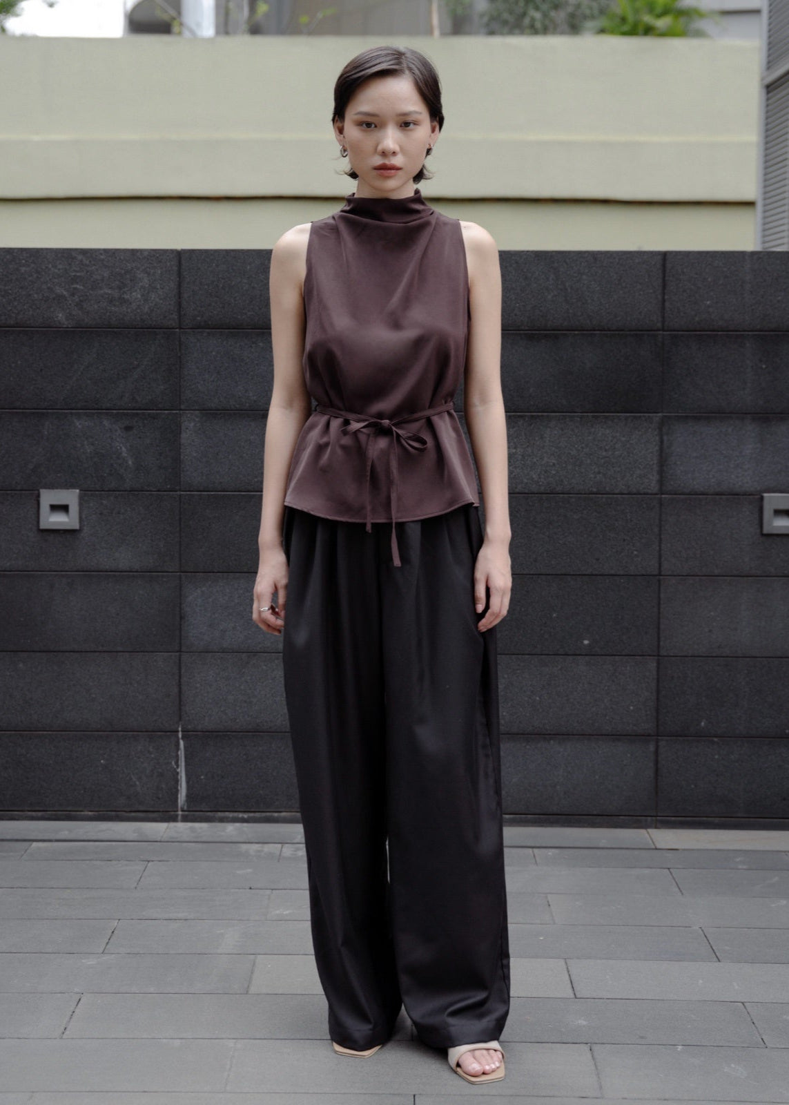 Woman wearing a sleeveless brown top and black pants standing against a dark stone wall.