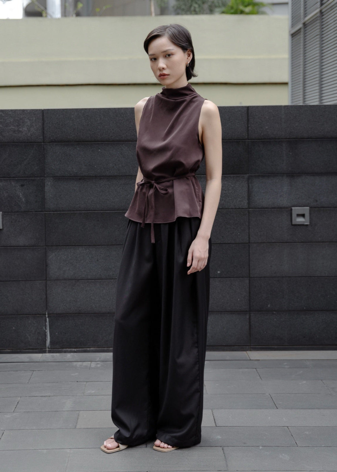 Woman wearing a sleeveless brown top and black pants standing against a gray wall.