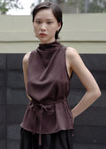 Woman wearing a brown sleeveless top with a high collar against a textured wall.