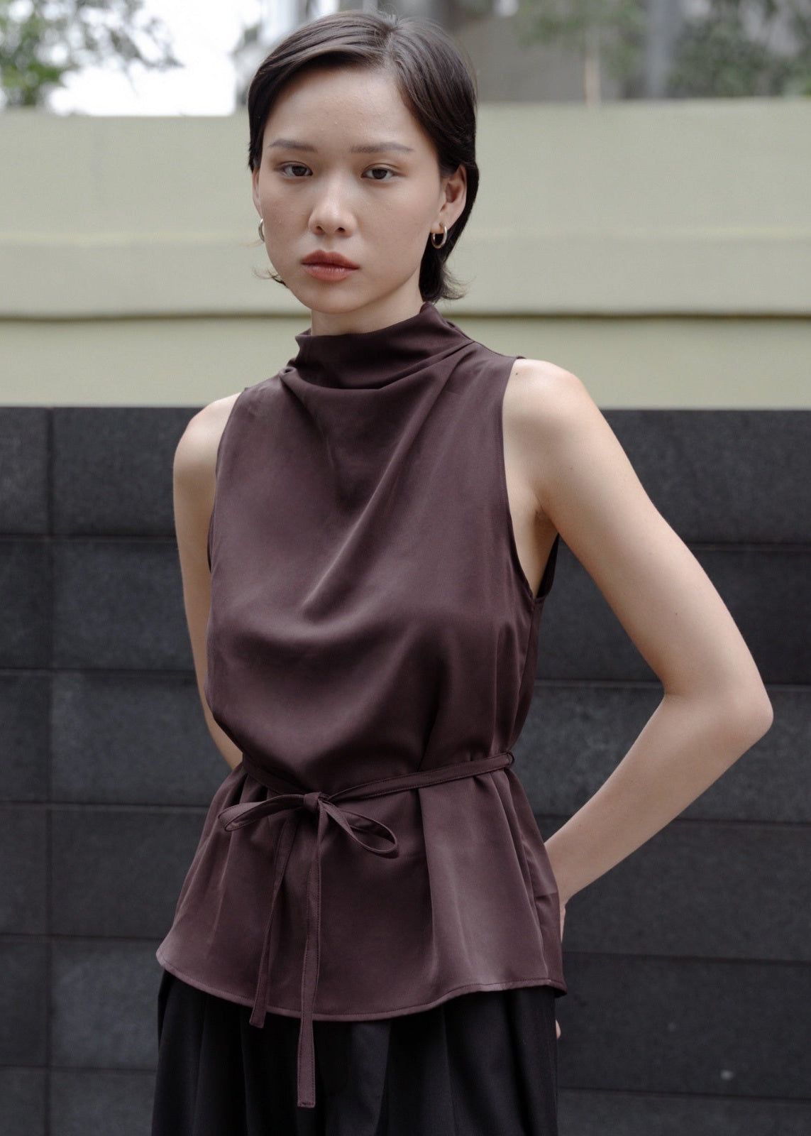 Woman wearing a brown sleeveless top with a high collar against a textured wall.