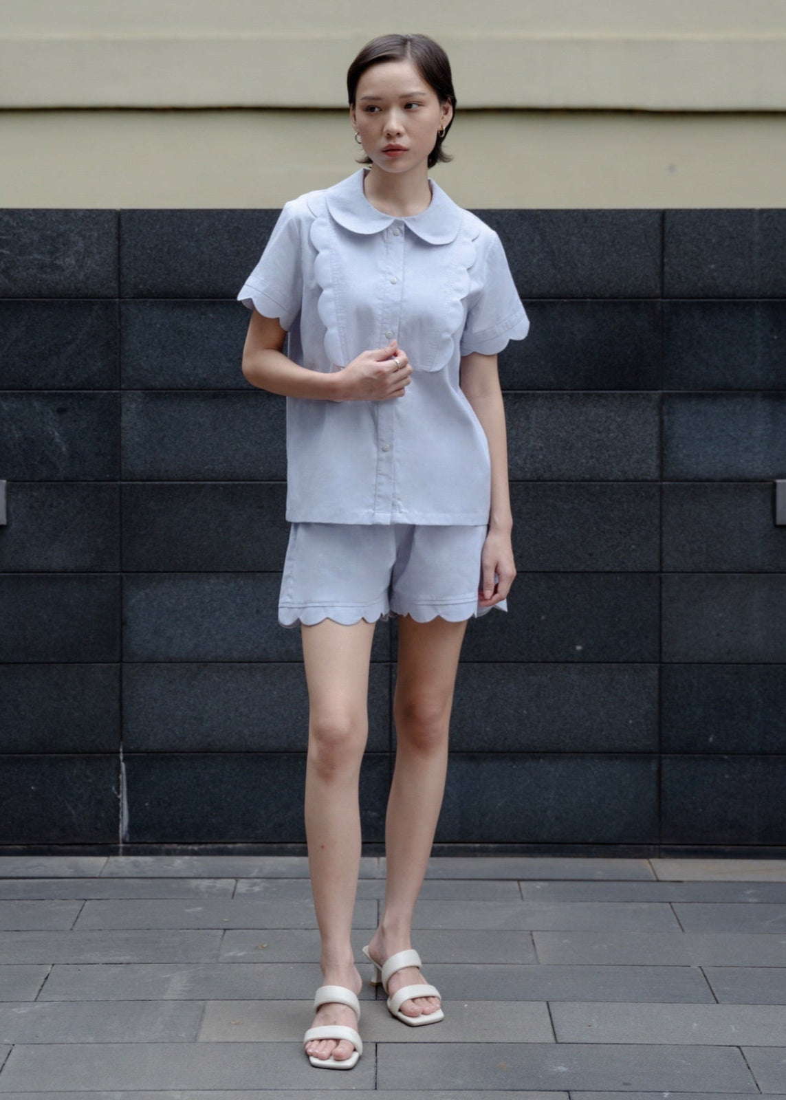 Woman wearing a light gray outfit standing against a dark tiled wall.