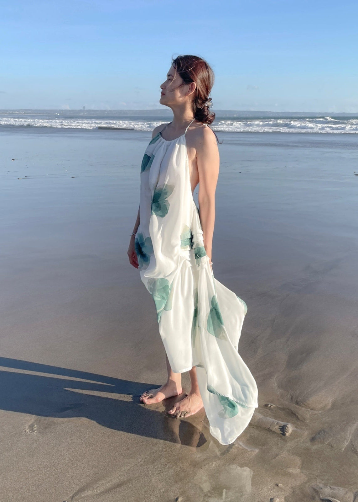 Woman in a white dress with green patterns standing on a beach.