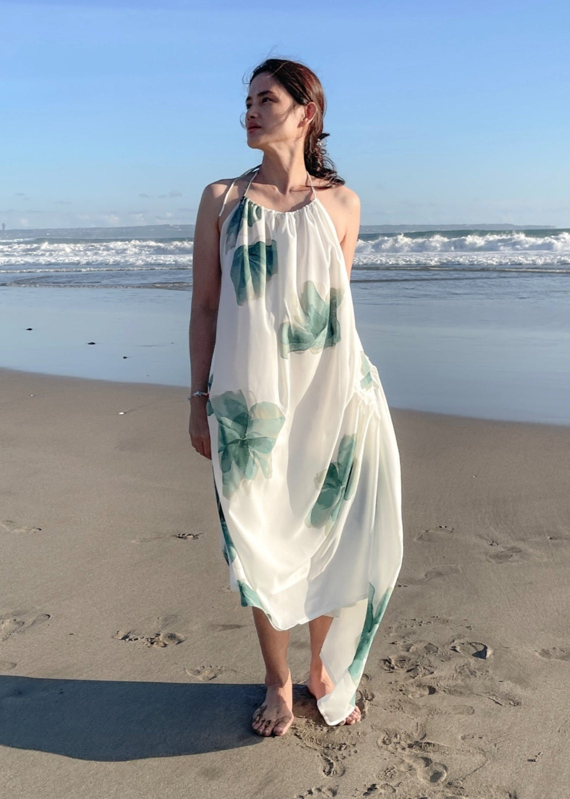 Woman in a white dress with green patterns standing on a beach.