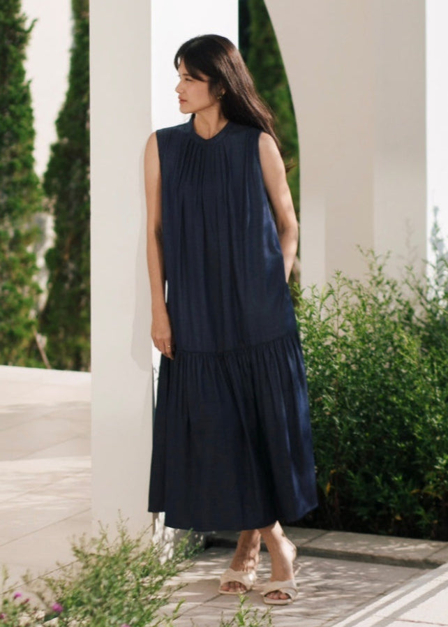 Woman wearing a navy blue dress standing outdoors with greenery and a white wall in the background