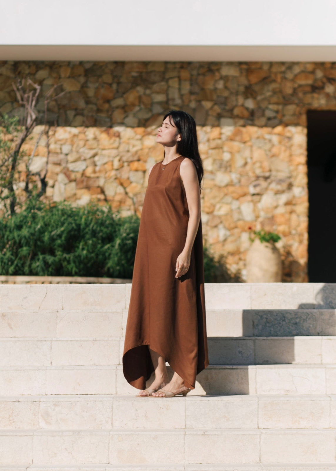 Woman in a brown dress standing on stone steps with a stone wall and greenery in the background
