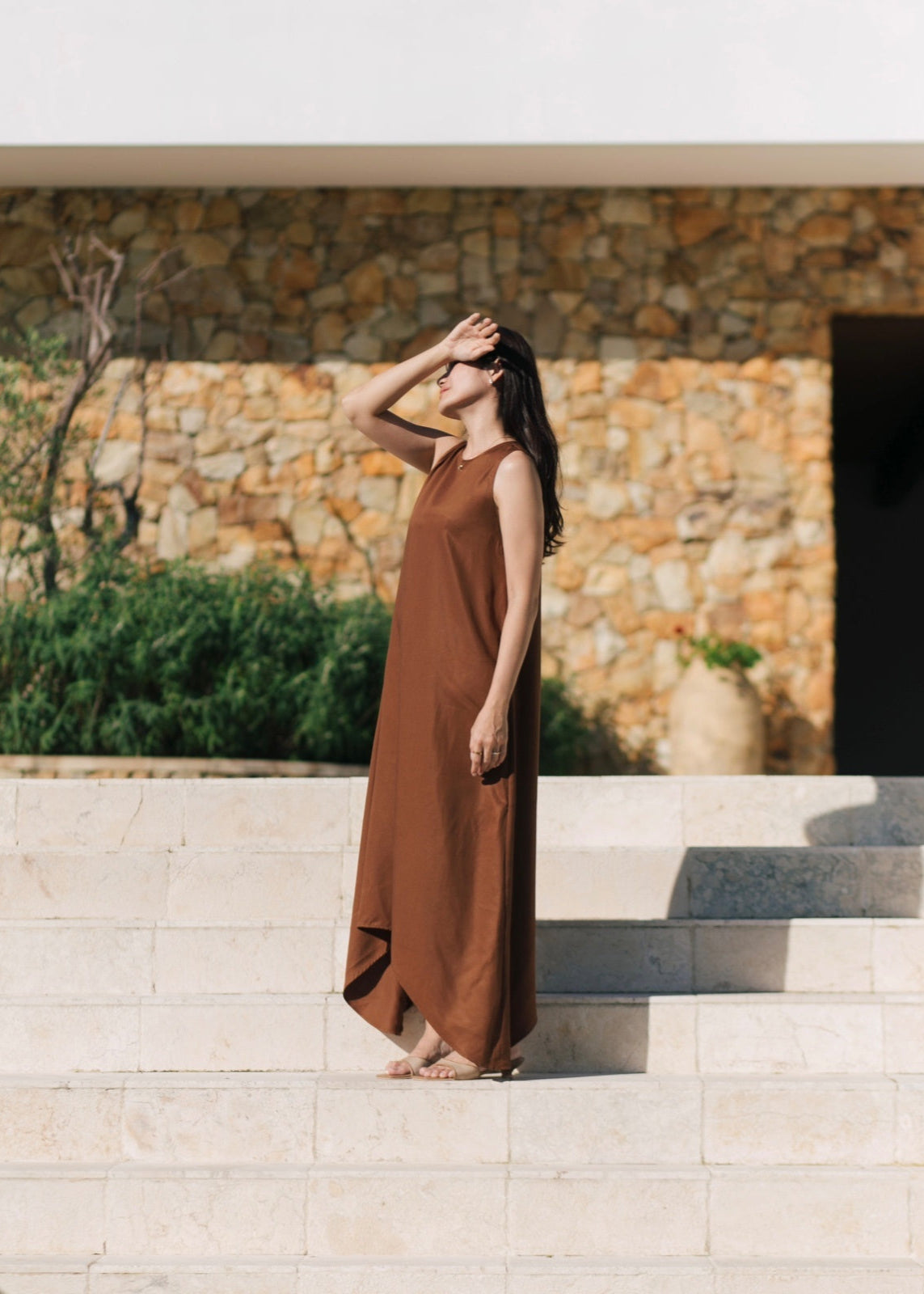 Woman in a brown dress standing on stone steps with a stone wall and greenery in the background