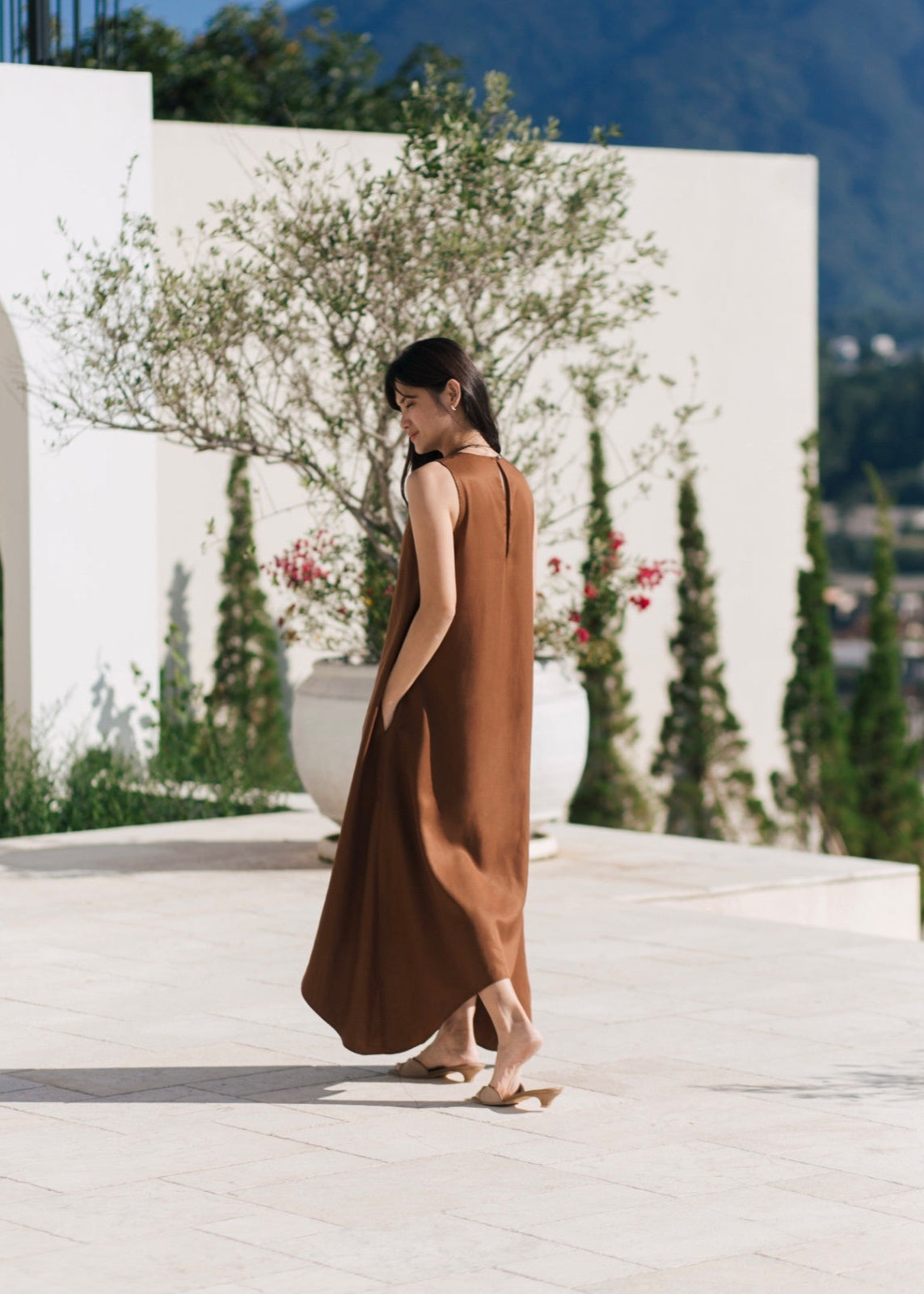 Woman in a brown dress standing in an outdoor setting with white architecture and greenery.