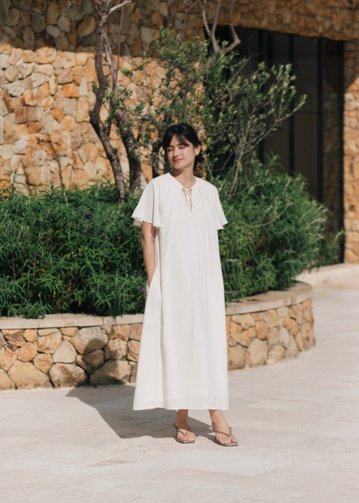 Woman in a white dress standing outdoors with stone wall and greenery in the background