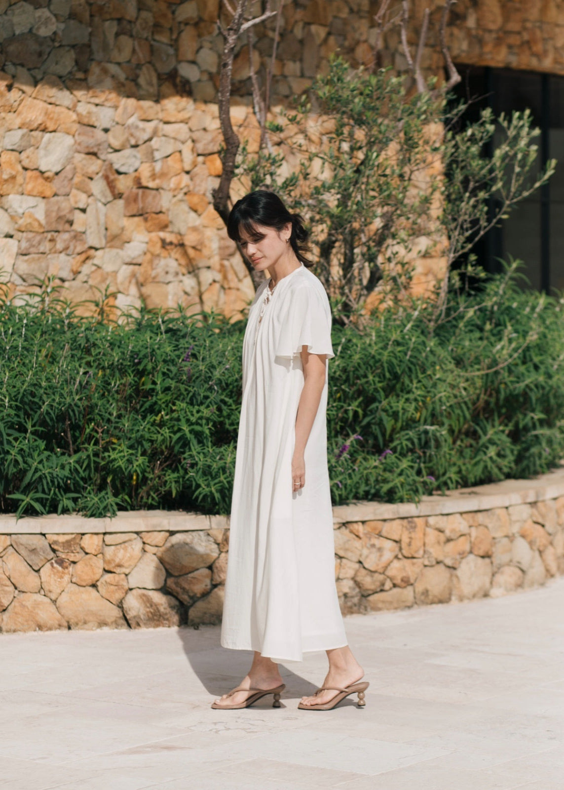 Woman in a white dress standing outdoors with a stone wall and greenery in the background