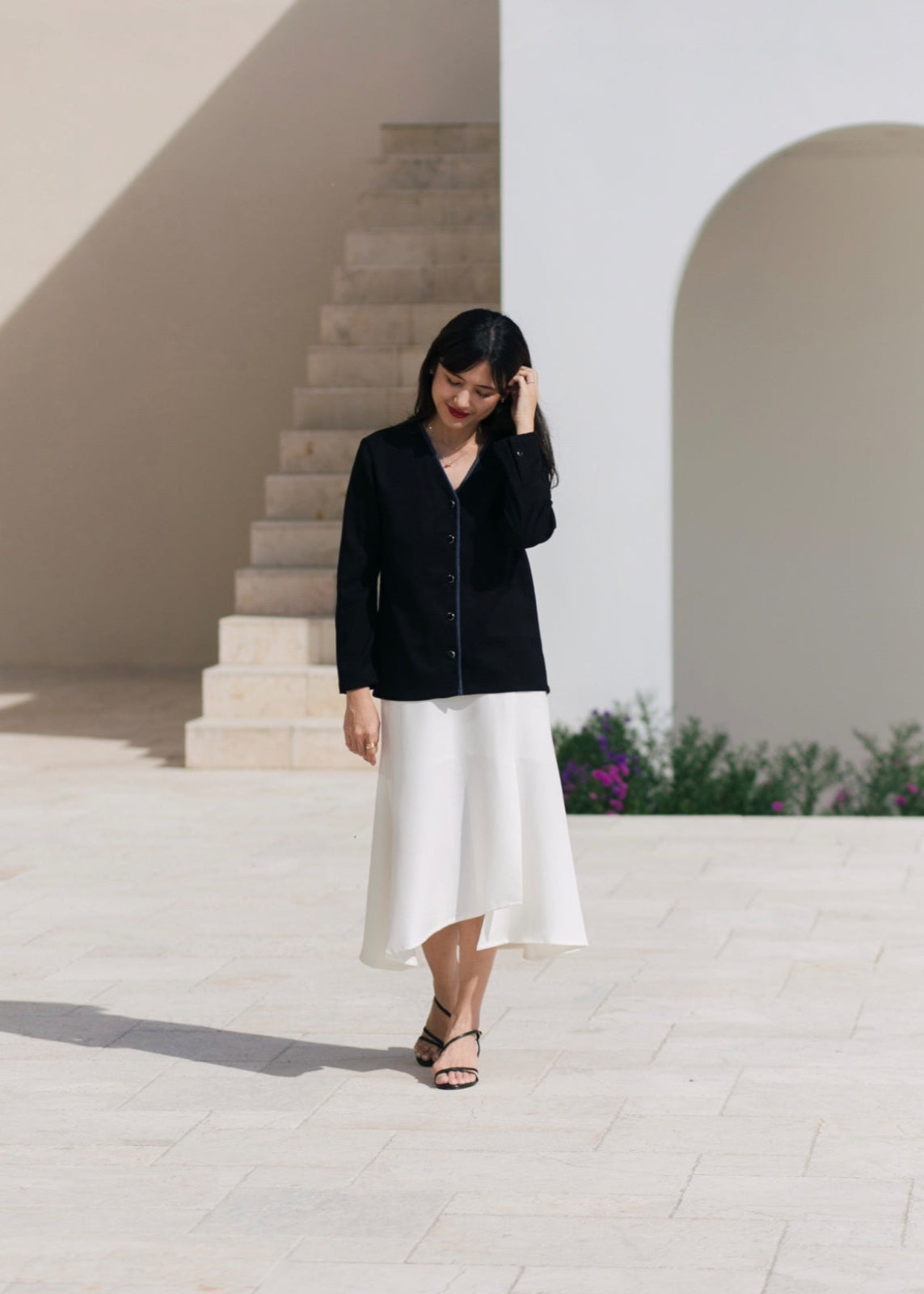 Woman in black shirt and white skirt standing in front of a white wall and steps.