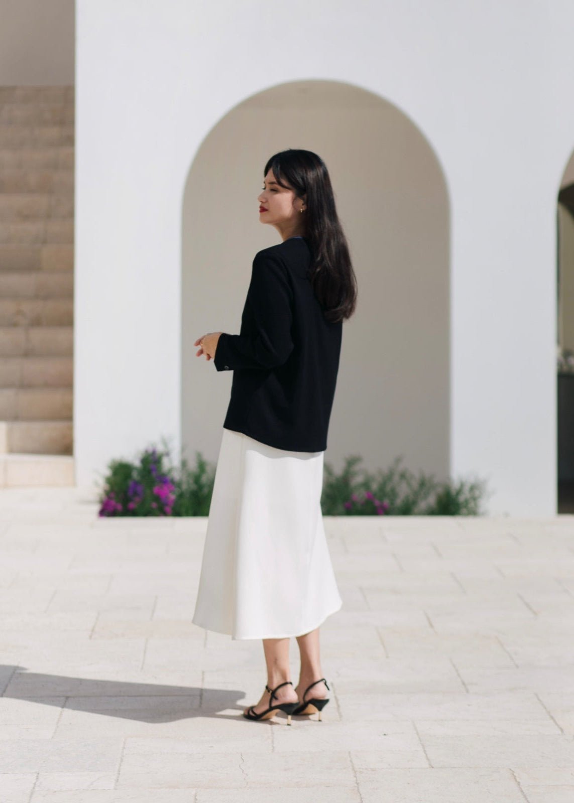 Woman in black blazer and white skirt standing in front of a white building with arches.