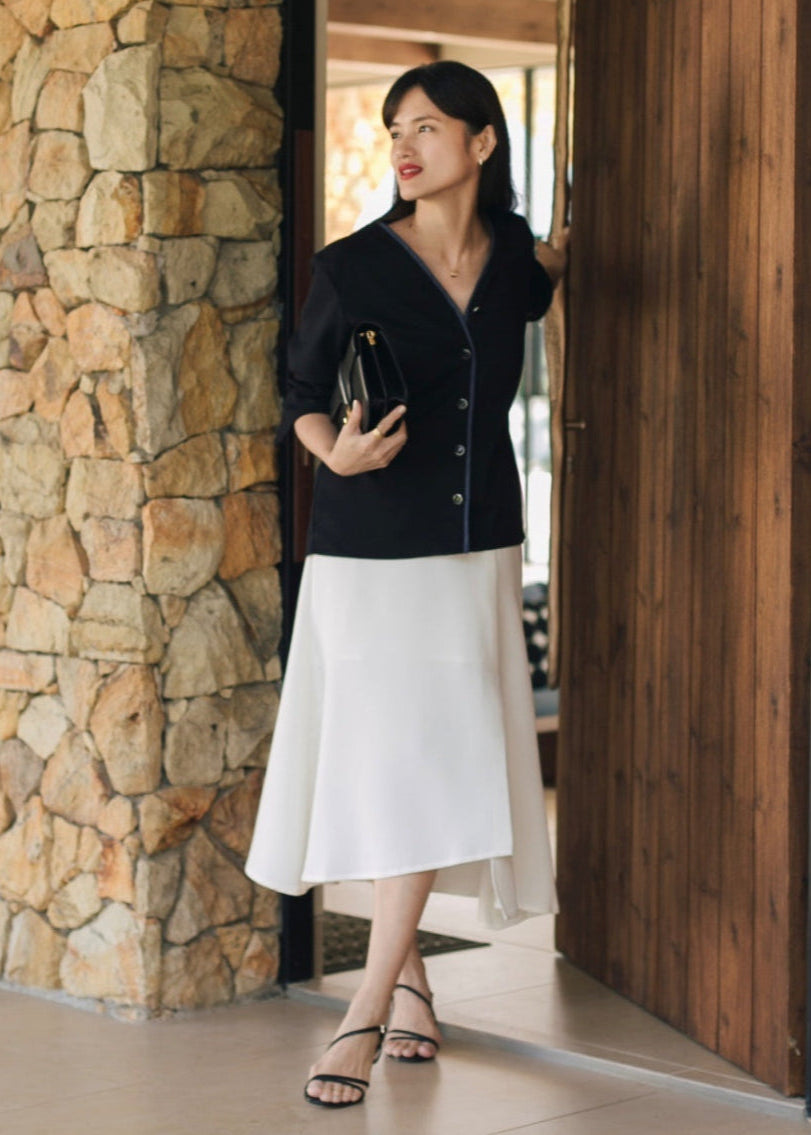 Woman in black jacket and white skirt standing in a modern interior setting with stone and wooden elements.